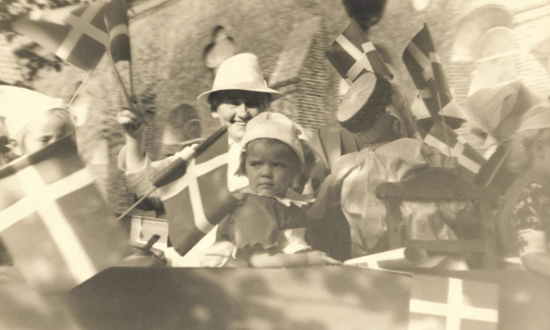 Kristine Kjeldsen (1890-1983), født i Vind og senere mangeårig forstanderinde ved Holstebro Børneasyl, ses siddende i baggrunden med hvid hat omgivet af børn fra asylet. Billedet er efter alt at dømme fra 1940’erne og taget enten i forbindelse med en børnehjælpsdag eller national festdag. I baggrunden genkendes Holstebro Sognekirkes høje mure.