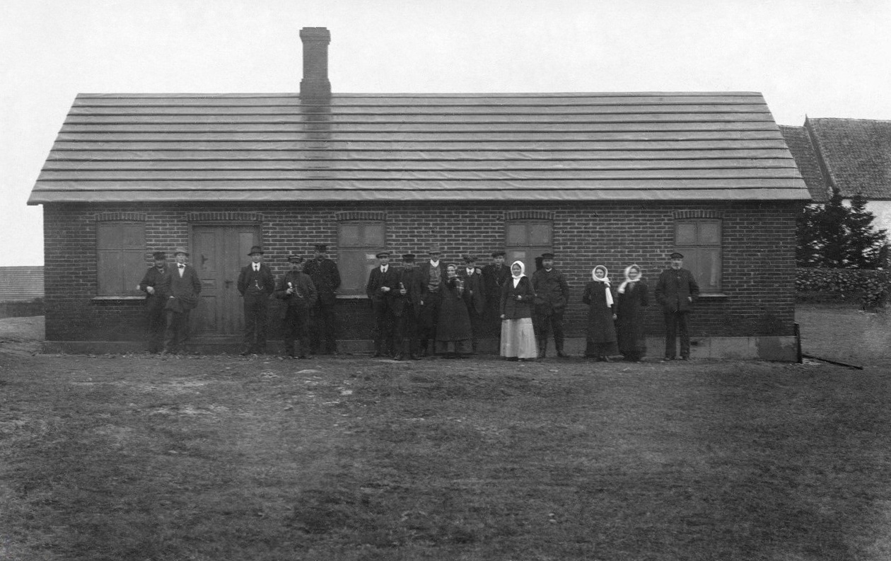 Forsamlingshuset, formentlig fotograferet omkring tidspunktet for dets indvielse i 1912. I baggrunden til højre genkendes kirken, og i baggrunden til venstre anes Toftegård. Enkelte af personerne lader sig identificere som bestyrelsens medlemmer, dog med en vis usikkerhed.